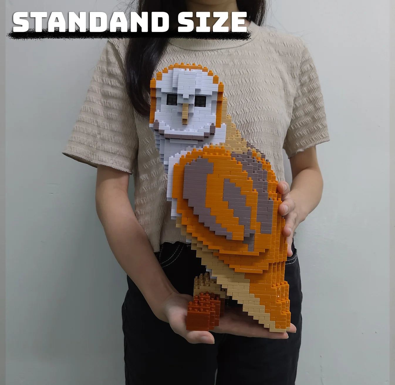 A person wearing a beige textured t-shirt holds a standard size, pixelated brick-built owl sculpture. The t-shirt features a graphic of an owl made from orange, white, and gray toy bricks.