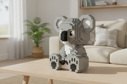 A pixelated gray koala figure made of small building blocks sits on a light wood table in a softly lit living room with a sofa, plant, and curtains in the background.