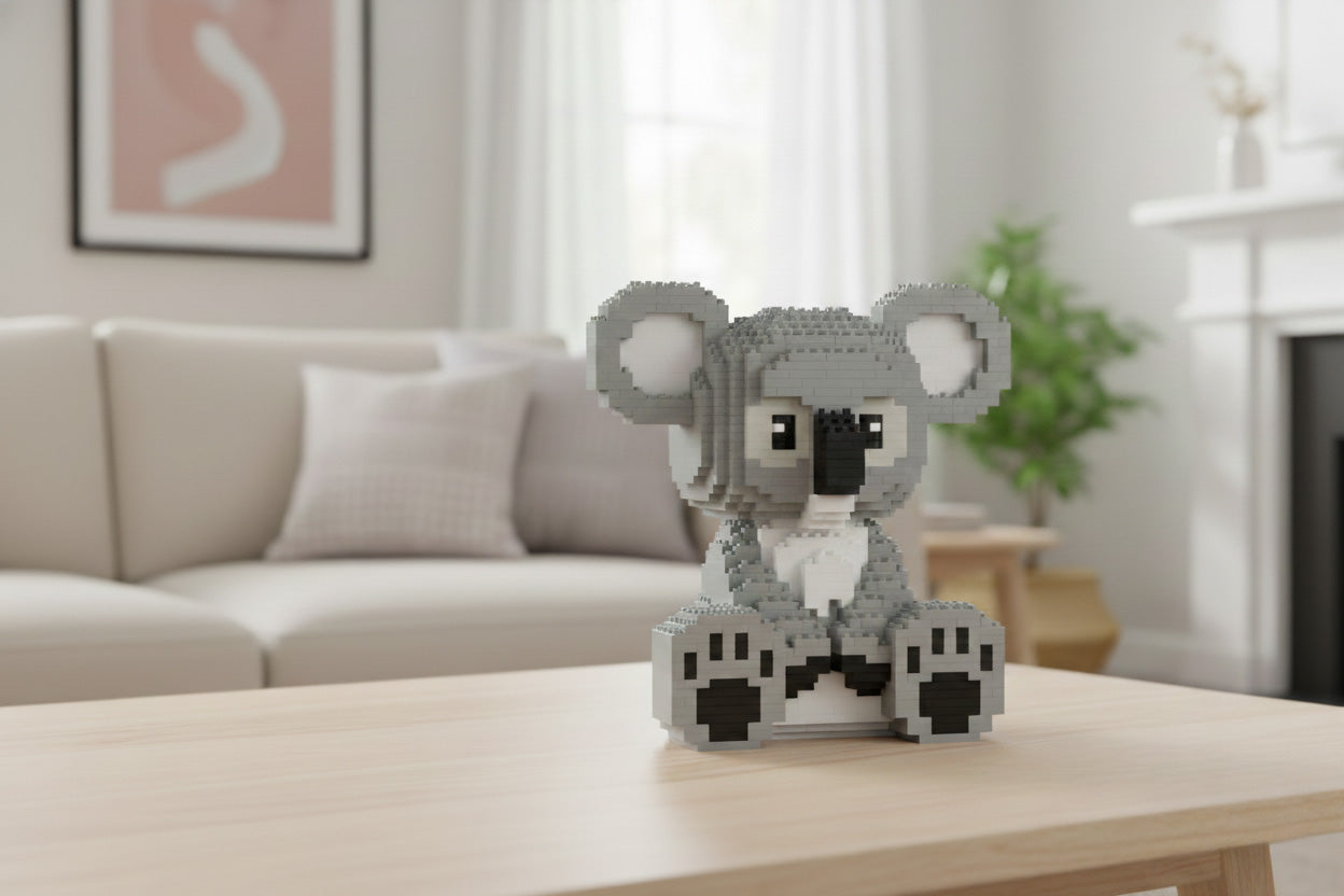A small gray koala figure made of interlocking blocks sits on a light wood table in a softly blurred living room with a sofa, framed wall art, and a plant in the background.