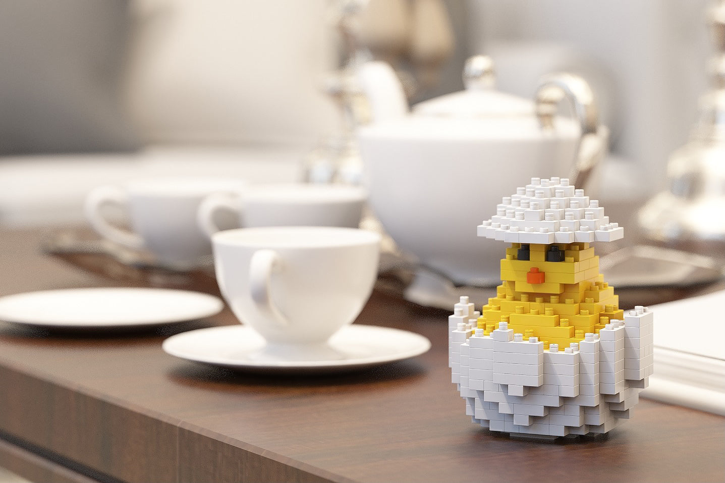 A small yellow chick toy built from interlocking blocks, wearing a white brick hat and sitting in a white brick egg, displayed on a wooden table in front of a blurred white teapot and teacups.
