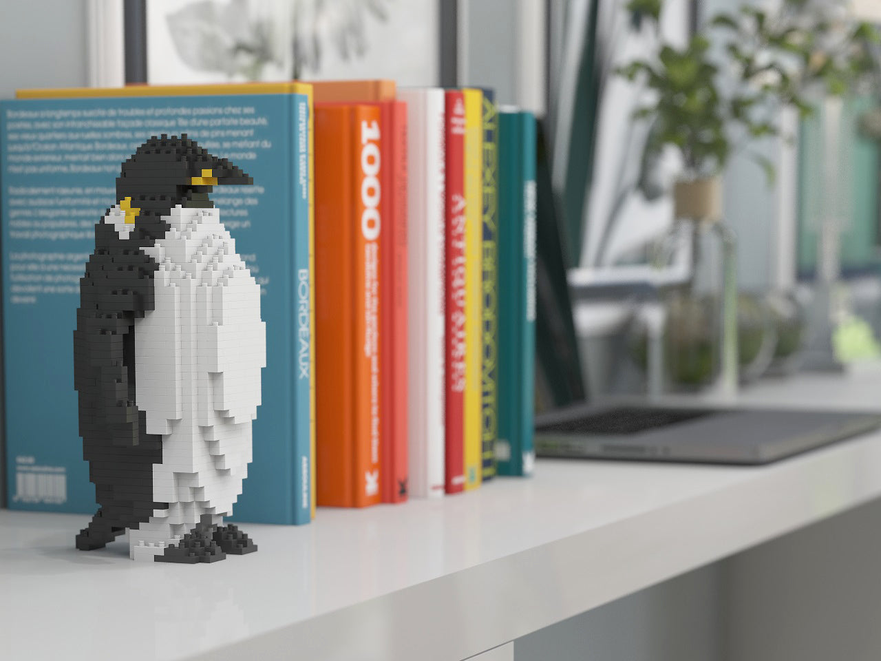 A penguin-shaped building block model made of small interlocking blocks stands on a white shelf in front of a row of colorful books, with a blurred indoor background including a plant and laptop.