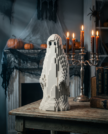 A white pixelated ghost-shaped decorative figure stands on a rustic wooden table in a Halloween-themed room, with lit candles, pumpkins, spider decorations, and old books in the background.