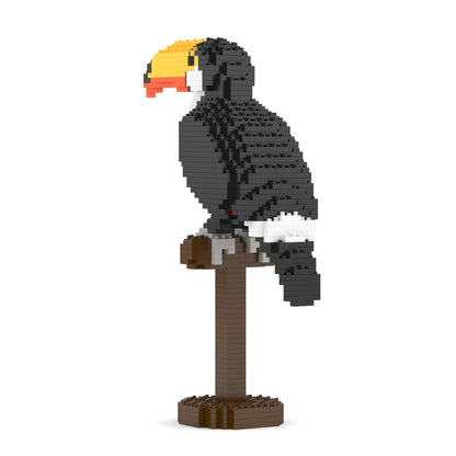 A toucan bird model built from small interlocking blocks, perched on a brown stand against a plain white background. The bird has a black body, white underparts, a yellow and orange beak, and gray details.